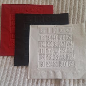 May include: Three bingo cards in red, black, and white. Each card has the word "BINGO" embossed at the top. The white card displays a grid of numbers, including 12, 19, 40, 59, and 65. The cards are on a white, textured surface.