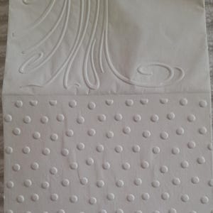 Pu&ograve; includere: Carta bianca strutturata con un design in rilievo. La parte superiore presenta linee curve e fluide, mentre la parte inferiore ha un motivo di cerchi in rilievo distanziati uniformemente. La carta sembra essere piegata.