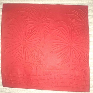 Könnte beinhalten: Ein rotes, rechteckiges Blatt mit einem geprägten Feuerwerksdesign. Das Design umfasst mehrere große, stilisierte Ausbrüche, kleine Sterne und einen strukturierten, ziegelartigen Rand am unteren Rand. Der Gesamteffekt ist festlich und feierlich.