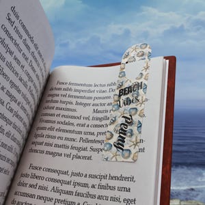 Puede incluir: Un marcapáginas decorativo con temática de playa, con conchas marinas y estrellas de mar. El marcapáginas tiene las palabras "Beach Vibes" y "Penny" impresas. Está insertado en un libro.