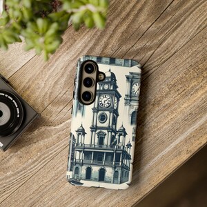 Peut inclure: Coque de téléphone avec une illustration architecturale d'une tour d'horloge en bleu et crème. Le design présente plusieurs cadrans d'horloge et des détails ornementaux. Posé sur une surface en bois.