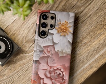 Può includere: Una custodia per telefono con un design floreale con fiori di carta rosa e bianchi. La custodia ha un ritaglio per la fotocamera con tre obiettivi.