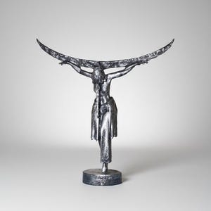 Op de afbeelding: Zilverkleurige sculptuur van een figuur met uitgestrekte armen die een groot, gebogen element ondersteunt. De figuur staat op een cirkelvormige basis, met een artistiek, abstract ontwerp.