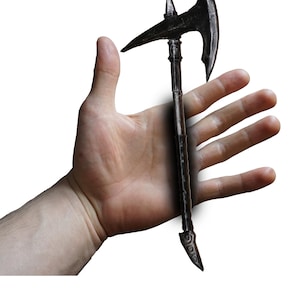 Puede incluir: Un hacha pequeña de estilo antiguo con un acabado metálico oscuro. El hacha presenta una hoja curva y una punta en el lado opuesto. El mango es largo y delgado, con elementos decorativos. El hacha se sostiene en una mano abierta sobre un fondo blanco.