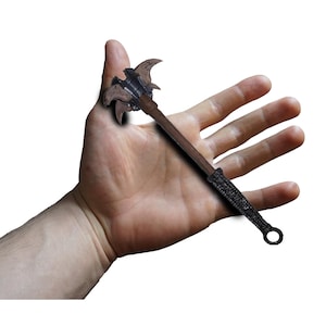 Puede incluir: Una maza medieval en miniatura con un mango de madera marrón oscuro y una cabeza texturizada detallada. La cabeza presenta múltiples cuchillas curvas y un acabado de metal oscuro. El mango tiene un agarre tejido y un anillo en el extremo.