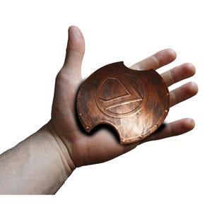 May include: A small, copper-colored shield with a stylized letter "L" in the center. The shield has a weathered appearance with a curved edge and rivet details. The shield is held in a hand against a white background.