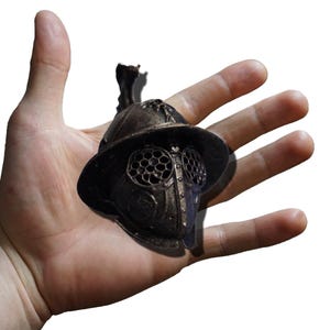 May include: A miniature bronze-colored gladiator helmet featuring a wide brim, a central crest, and a protective face mask with circular mesh eye coverings. The helmet has a weathered, metallic finish with visible rivets and structural detailing.
