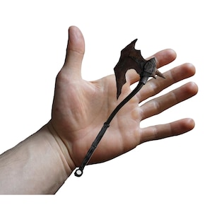 May include: A miniature, rusty-brown axe with a jagged blade and a long, slender handle. The axe is held in an open hand against a white background, showcasing its small size and detailed craftsmanship. The axe appears to be made of metal.