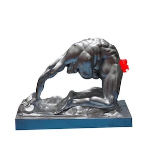 Può includere: Una scultura color argento di una figura inginocchiata, che si piega all'indietro. La figura è molto dettagliata, con muscolatura visibile. La scultura è montata su una base rettangolare blu scuro, con una superficie strutturata.