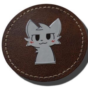 May include: A circular brown leather coaster featuring a white, stylized cartoon cat character with black outlines and small red markings on its cheeks. The coaster has visible light-colored stitching along the perimeter of the circular edge.