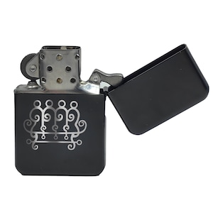 May include: A black metal lighter with a silver decorative design and a hinged lid. The lighter is open, revealing the interior. The design is a series of connected, stylized figures. The lighter's top is silver with a wheel and a flint.