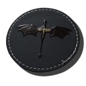 May include: A circular black patch featuring a debossed dragon silhouette in the centre with outstretched wings and a spiked tail. The patch has a visible white stitched border around the entire circumference, creating a distinct contrast against the dark material.