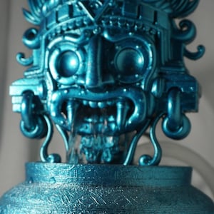 May include: A metallic blue fountain featuring a sculpted deity mask with prominent fangs and an elaborate headdress. The base is a rounded vessel with intricate, swirling embossed patterns. Water flows from the mouth of the central figure.