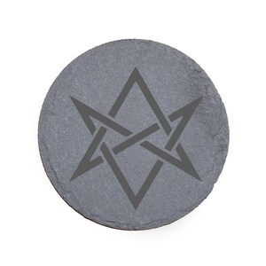 Pode incluir: Uma base redonda de ardósia cinza com um desenho de estrela escura entrelaçada. A base tem uma superfície natural e texturizada e uma borda ligeiramente irregular. O desenho está centrado.