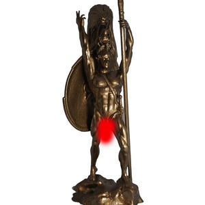 Puede incluir: Estatua de color bronce de una figura musculosa que sostiene una lanza y un escudo. La figura está de pie sobre una base rocosa con un brazo levantado. Una cabeza de león está detrás de la figura.