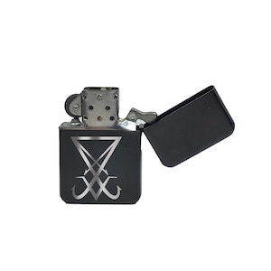 May include: Black metal lighter with a silver emblem. The lighter is open, revealing the interior. The emblem is a stylized, inverted cross with two curved lines. The lighter has a silver hinge and a silver top.