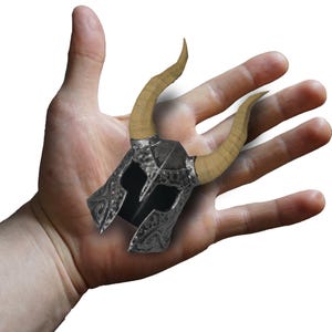 May include: A miniature metallic helmet featuring a dark, hammered texture and two prominent, curved tan horns extending from the sides. The helmet has a central nasal guard and decorative scrollwork patterns etched along the lower edges.