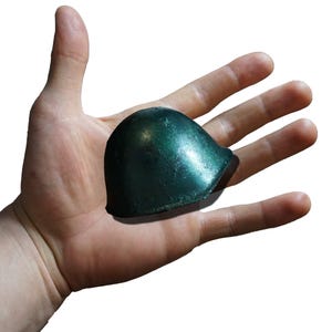 May include: A miniature, dome-shaped helmet with a smooth, metallic dark green finish. The object features a curved brim and a rounded top, reflecting light across its surface. This small, solid-looking piece is presented against a plain white background.