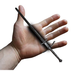 Peut inclure: Une épée gris foncé avec une garde transversale et une pointe. L'épée est tenue dans une main ouverte sur fond blanc. La poignée de l'épée a un motif texturé.