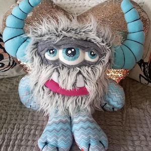 May include: A plush gray monster toy with a single blue glitter eye, a pink mouth with teeth, and two blue horns. The monster is sitting on a gold heart-shaped pillow with a blue and gray chevron pattern on its legs.