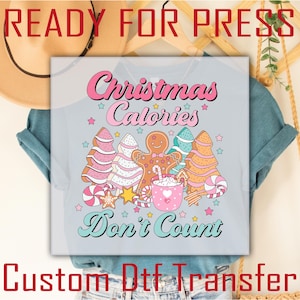 Puede incluir: Camiseta azul claro con un gráfico navideño colorido. El diseño presenta hombrecitos de jengibre, árboles de Navidad, bastones de caramelo y el texto "Christmas Calories Don't Count" en rosa y turquesa.