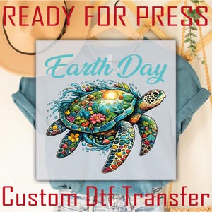 May include: Light blue t-shirt featuring a colorful sea turtle design with the text "Earth Day". The turtle's shell is filled with flowers and a sun, surrounded by water. The words "READY FOR PRESS" and "Custom Dtf Transfer" are also visible.