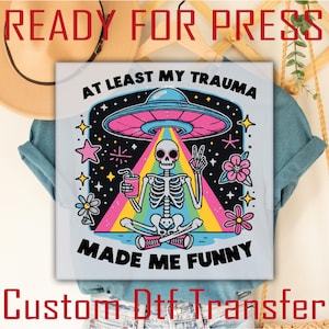 May include: A white t-shirt with a colorful graphic design featuring a skeleton sitting in a lotus position with a UFO above. The text "At least my trauma made me funny" is printed on the shirt.