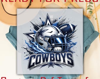 May include: A light gray t-shirt featuring a graphic print of a silver football helmet with a blue face mask, a football, and a large star. The text COWBOYS is printed in bold blue letters below the helmet, surrounded by blue and black ink splashes.