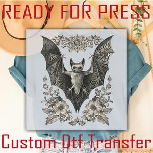May include: A light blue t-shirt featuring a detailed black and white illustration of a bat with outstretched wings, surrounded by floral elements. The text "READY FOR PRESS" and "Custom Dtf Transfer" are printed in red.