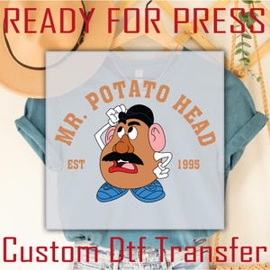 May include: A light blue t-shirt with a custom iron-on transfer design featuring Mr. Potato Head. The design includes the text "Mr. Potato Head" in a brown circle with the text "Est 1995" and a cartoon image of Mr. Potato Head.