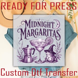May include: A light blue t-shirt featuring a purple graphic print with the text Midnight Margaritas. The design includes a margarita glass, a moka pot, a frog wearing a witch hat, a crescent moon, and sprigs of lavender in a detailed illustration.