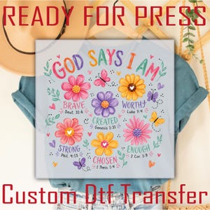 May include: A light blue t-shirt featuring a colorful graphic print with the text "GOD SAYS I AM" surrounded by various flowers, butterflies, and small hearts. The design includes inspirational words like "BRAVE," "WORTHY," "CREATED," "STRONG," "ENOUGH," and "CHOSEN" paired with Bible verse references.