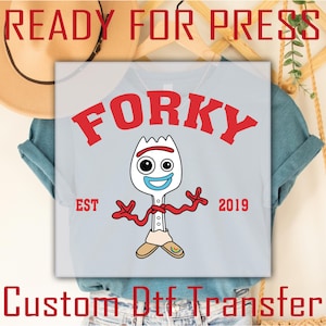 May include: A light blue t-shirt with a red and white graphic of the character Forky from the movie Toy Story. The graphic includes the text "Forky" in red, "EST" and "2019" in black, and a rainbow on Forky's shoe. The text "Custom Dtf Transfer" is printed in red at the bottom of the image.