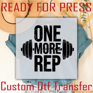 May include: A light blue t-shirt with the text "ONE MORE REP" in bold black letters, flanked by barbell illustrations. The words "READY FOR PRESS" and "Custom Dtf Transfer" are also visible in red and black.