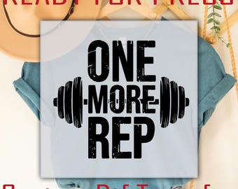 Könnte beinhalten: Hellblaues T-Shirt mit dem Text "ONE MORE REP" in schwarzer Schrift, flankiert von Hantel-Illustrationen. Die Wörter "READY FOR PRESS" und "Custom Dtf Transfer" sind ebenfalls in Rot und Schwarz zu sehen.