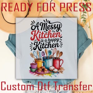 May include: A light blue t-shirt featuring a graphic print with the text A Messy Kitchen is a happy Kitchen in black and red script. The design includes three colorful mugs filled with various wooden and metal kitchen utensils and baked goods.