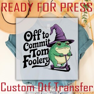 May include: A light blue t-shirt with a custom iron-on transfer design. The design features a cartoon frog wearing a purple witch hat and riding a scooter. The text "Off to Commit Tom Foolery" is printed in black.