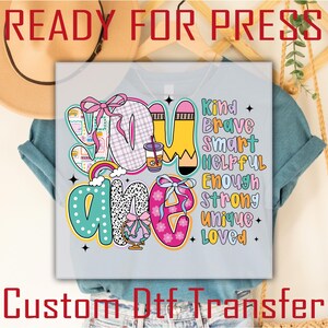 May include: A light gray t-shirt with a colorful graphic that reads "YOU ARE" with the words "Kind, Brave, Smart, Helpful, Enough, Strong, Unique, Loved". The words are in various colors and fonts. The shirt also has the text "READY FOR PRESS" and "Custom Dtf Transfer".