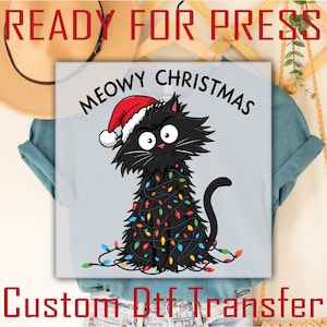 May include: A light blue t-shirt featuring a black cat decorated as a Christmas tree, wearing a Santa hat. The cat is adorned with colorful Christmas lights. Text above reads "MEOWY CHRISTMAS". Below the image is the text "Custom Dtf Transfer".
