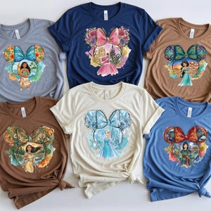 May include: A collection of six t-shirts in various colors, each featuring a unique graphic design. The designs incorporate a bow-shaped frame with Disney princess illustrations. The shirts are in shades of blue, brown, tan, and grey.