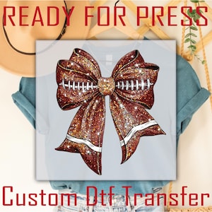 May include: A brown glitter bow with football stitching on a light blue t-shirt. The shirt is labeled "Custom Dtf Transfer" and "Ready For Press".