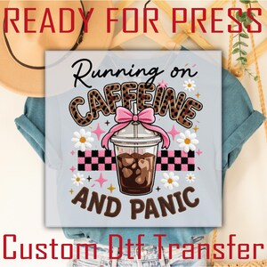May include: A light blue t-shirt with a graphic that reads "Running on Caffeine AND PANIC." The word "Caffeine" is in a leopard print design. The graphic includes a coffee drink, daisies, and a pink bow. The text "READY FOR PRESS" and "Custom Dtf Transfer" are also visible.