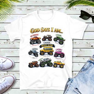 Puede incluir: Camiseta blanca con un gr&aacute;fico colorido de monster trucks y el texto "GOD SAYS I AM..." junto con afirmaciones positivas. El dise&ntilde;o incluye frases como "Victorioso", "Elegido" y "Fuerte", con vers&iacute;culos b&iacute;blicos.