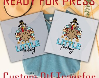 May include: Two square DTF transfers featuring a cartoon turkey wearing a black pilgrim hat, with colorful patterned tail feathers and small pumpkins at its feet. The text reads Little Turkey in a combination of light blue block and black script fonts.