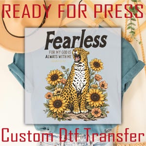May include: A light blue t-shirt featuring a graphic of a leopard surrounded by sunflowers. The text "Fearless" is above the leopard, with the phrase "For my God is always with me" below. The words "Ready for Press" and "Custom Dtf Transfer" are also visible.