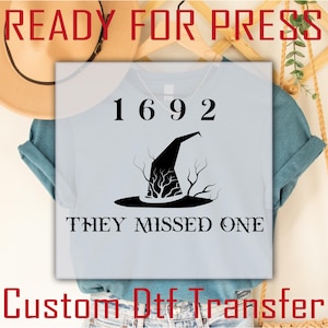 May include: Light blue t-shirt with the text "1692 THEY MISSED ONE" and a black witch hat graphic. The words "READY FOR PRESS" and "Custom Dtf Transfer" are also visible. The shirt is displayed on a hanger.