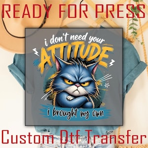 May include: A grey t-shirt featuring a graphic of a grumpy blue cat with crossed arms. The text reads i don't need your ATTITUDE i brought my own in yellow and black lettering. The design includes stylized lightning bolts and star accents.