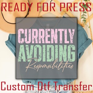 May include: A dark gray t-shirt featuring the text "CURRENTLY AVOIDING Responsibilities" in a distressed font. The word "CURRENTLY" is in light pink, "AVOIDING" is in light green, and "Responsibilities" is in a thin, cursive light pink script.