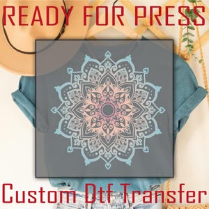 May include: A gray t-shirt with a blue and pink mandala design. The design is ready to be pressed onto a shirt using a heat press. "Custom Dtf Transfer" is written in red at the bottom of the image.
