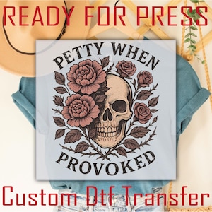 May include: A light blue t-shirt featuring a graphic print of a human skull partially surrounded by dark pink roses and leaves. The text PETTY WHEN PROVOKED is printed in a black arched font above and below the floral skull illustration.